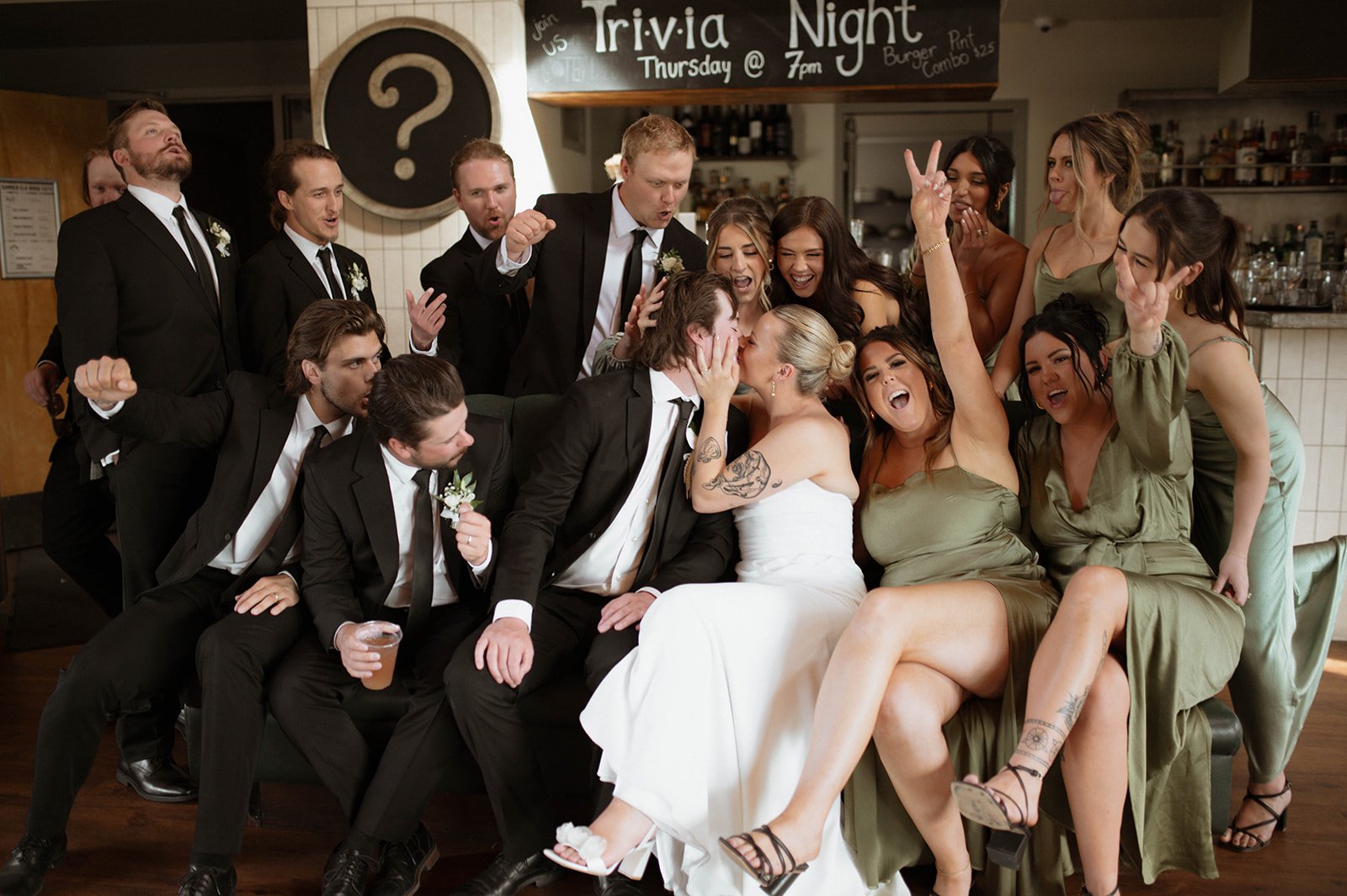 A group of wedding guests and the bride and groom gather around and kiss in a celebratory moment.