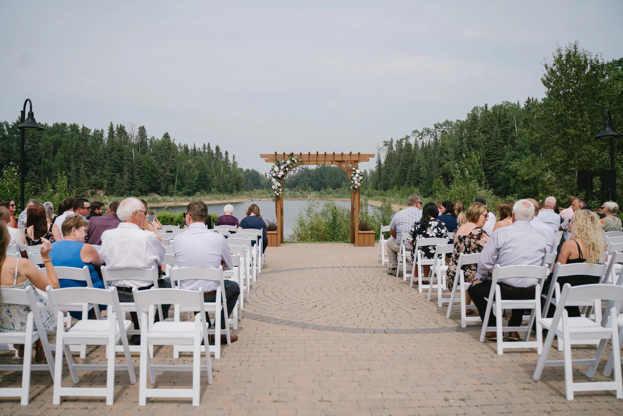 An outdoor wedding ceremony by a lake with a wooden arch decorated with flowers.
