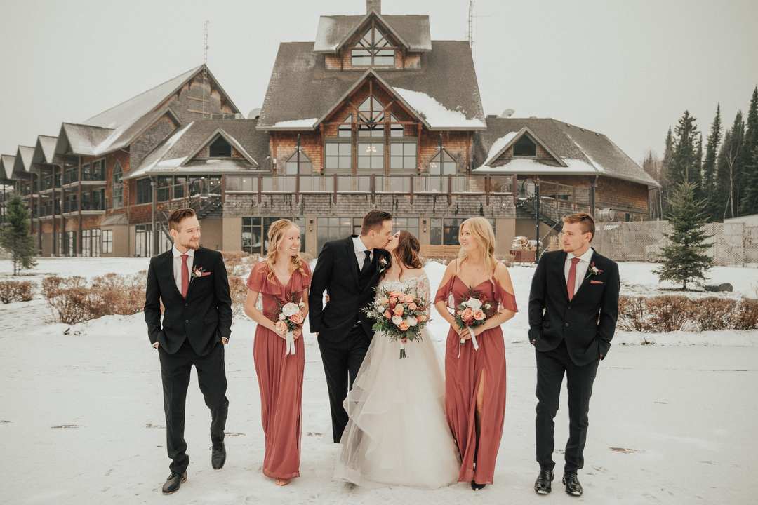 Wedding party standing on snowy ground in front of a large house, with two men in dark suits.