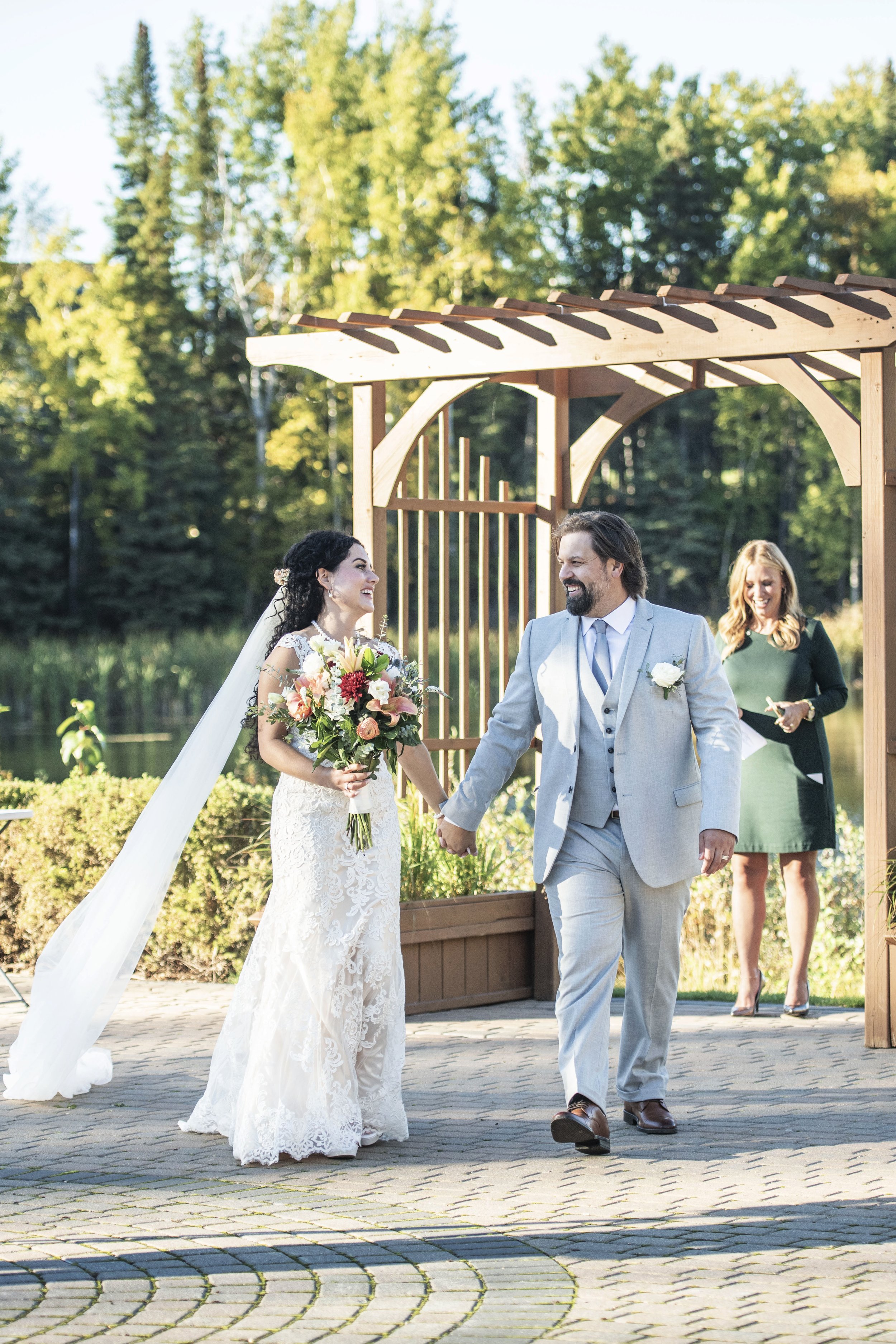 A bride and groom holding hands during their outdoor wedding ceremony, with a wooden arch behind them.