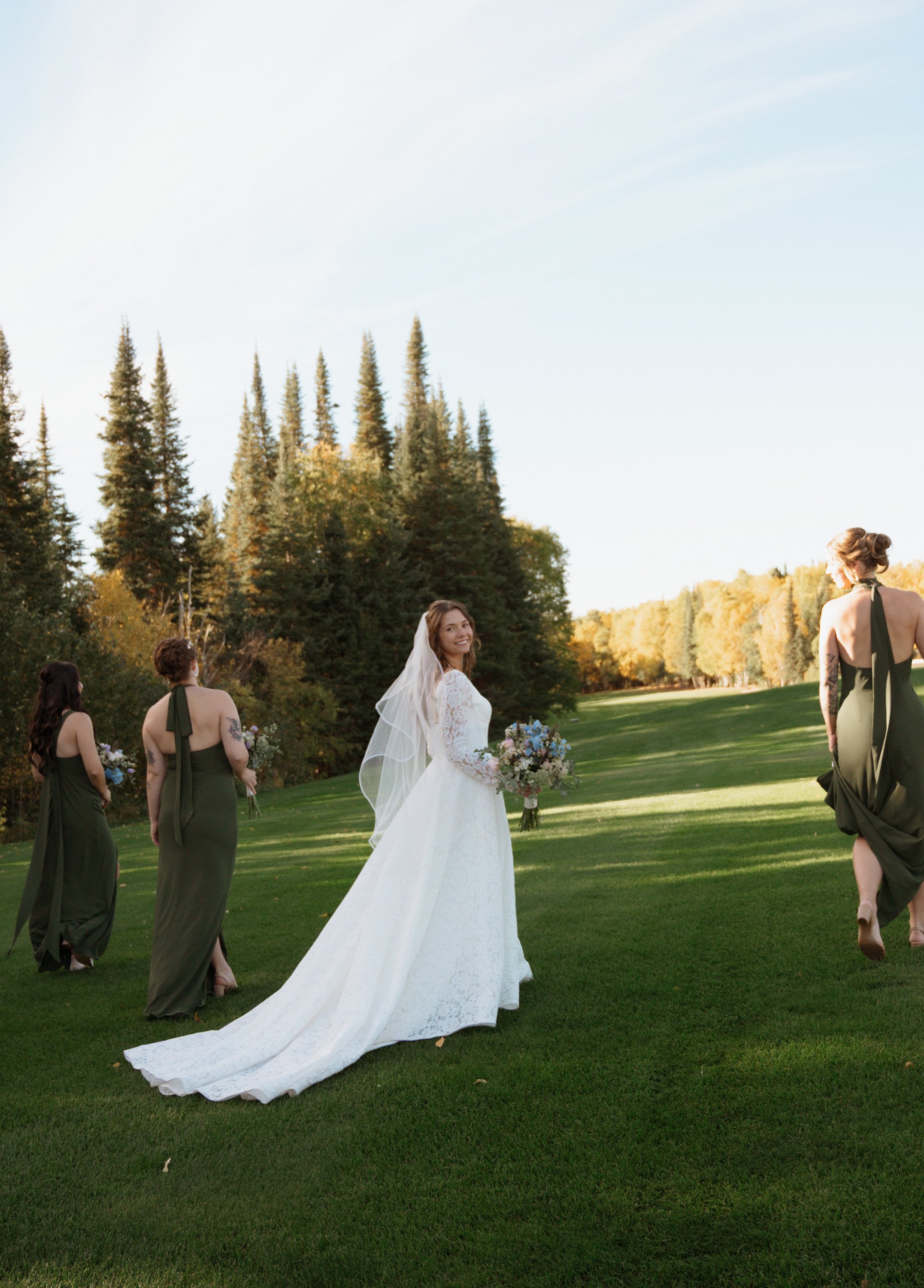 A bride in a white wedding gown stands outdoors on a grassy field, surrounded by bridesmaids.