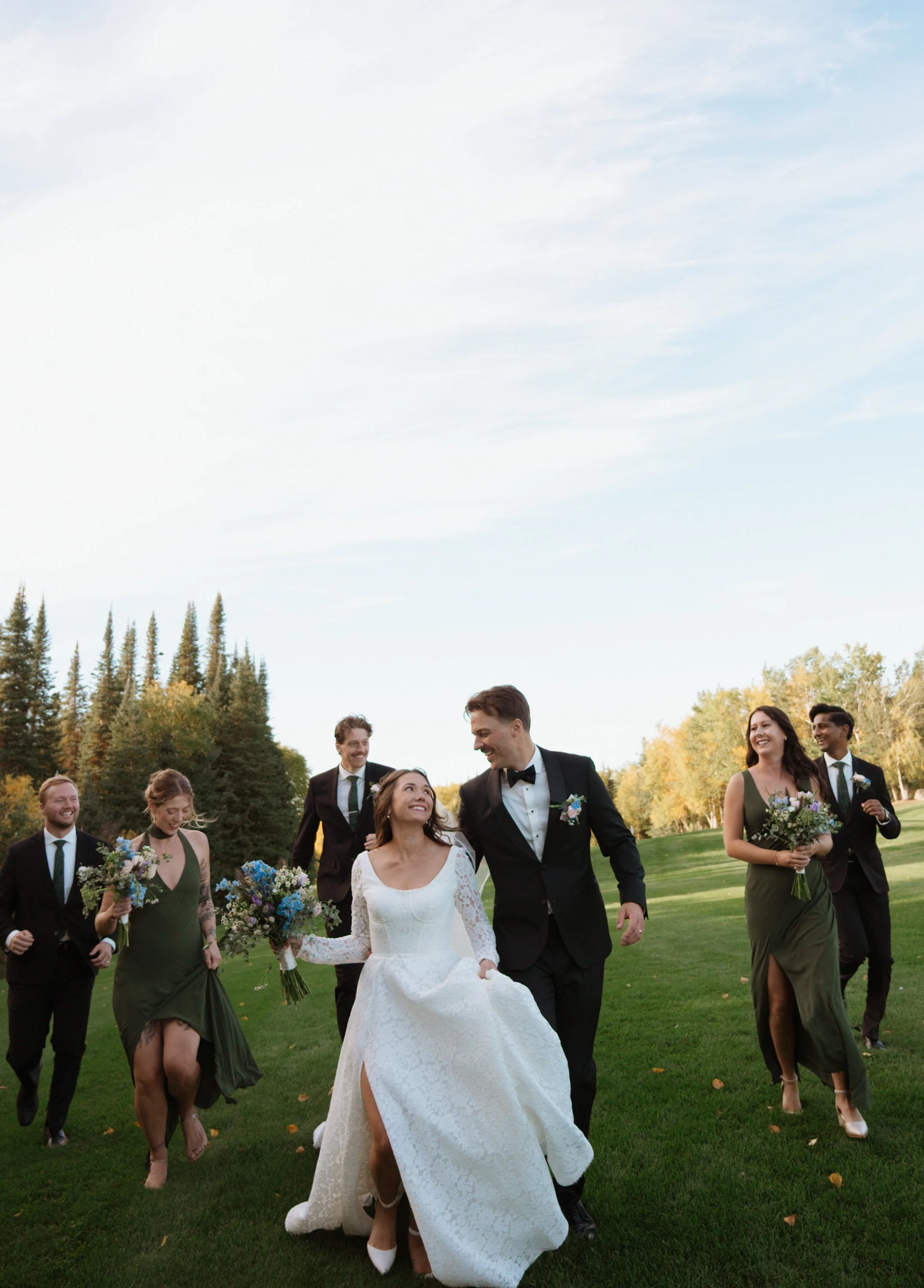 A wedding party, including the bride in a white dress and groom in a black tuxedo, walking on green grass.