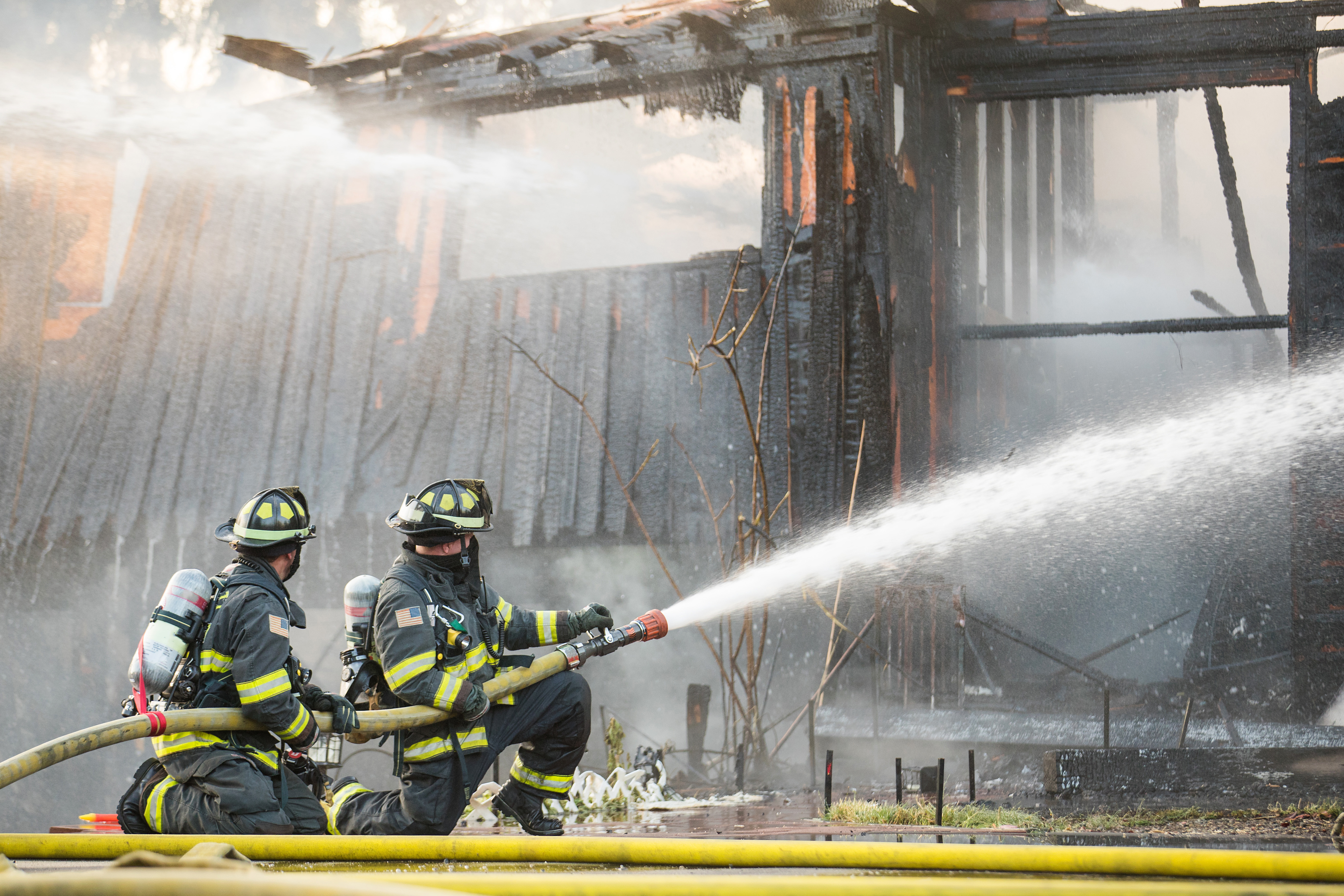 Two firefighters kneeling with a hose, spraying water onto a burned-out building