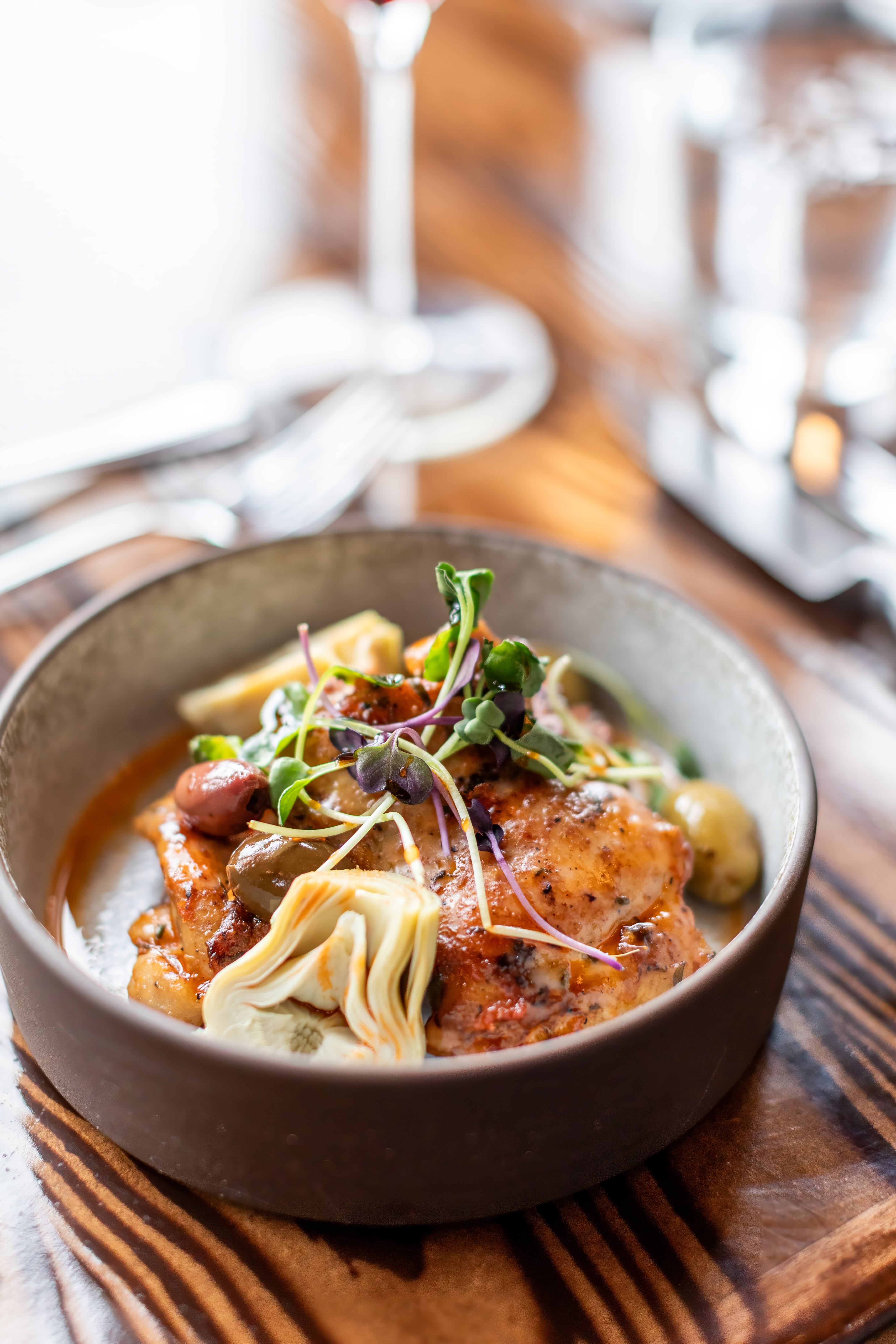 A plated chicken dish with artichoke, olives and microgreens in a stoneware bowl on a wooden board