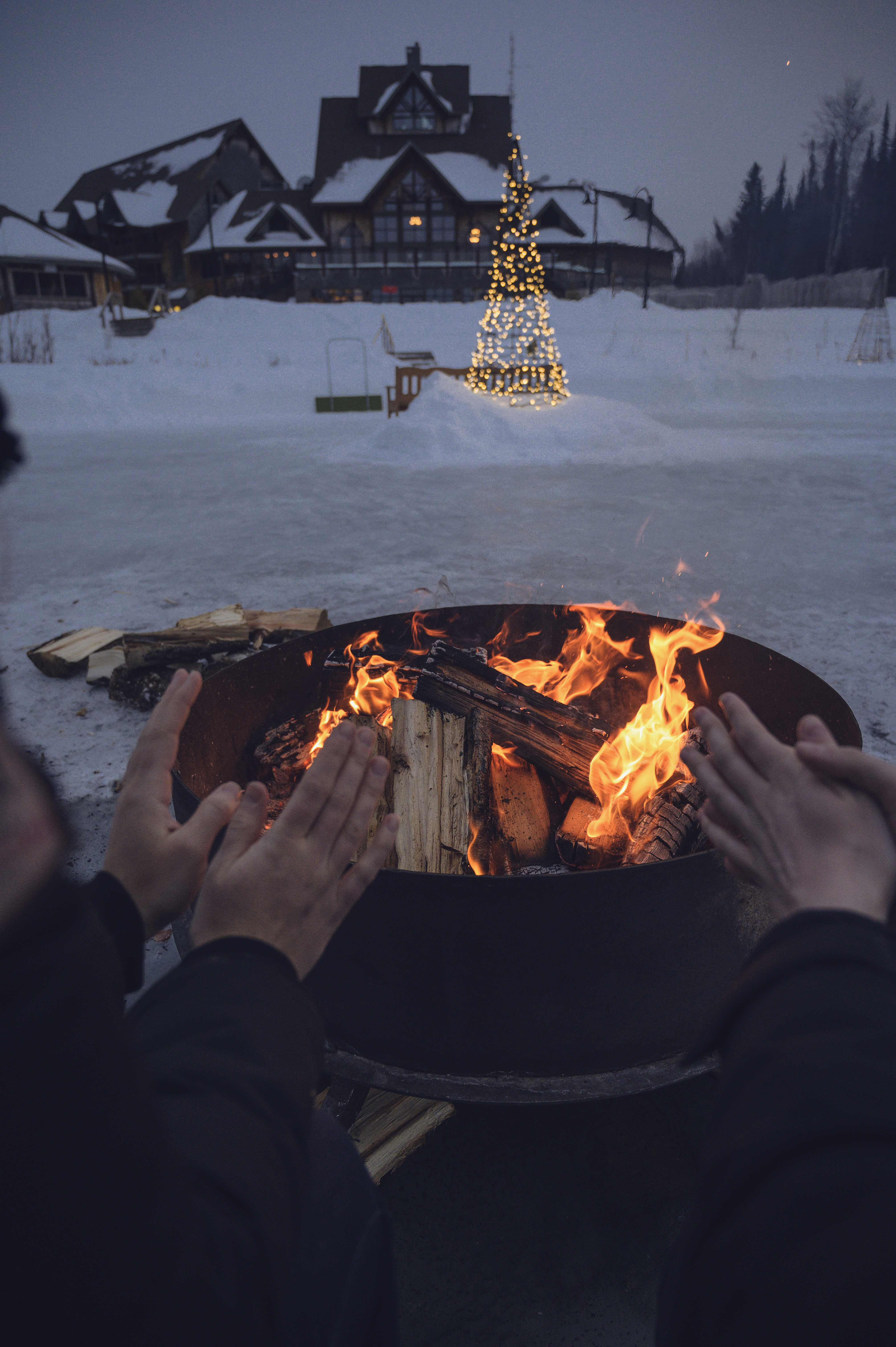 Hands warming at a fire pit on the snow, with the Lodge and a lit Christmas tree behind
